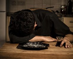 Sad and lonely man is resting his head on his dinner table in his kitchen and clutching a glass of wine